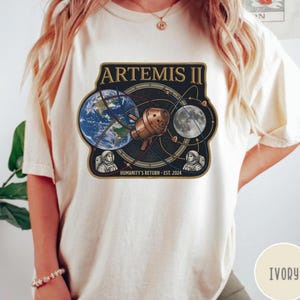 May include: Ivory-colored t-shirt featuring a graphic of the Artemis II mission. The design includes Earth, the Moon, a spacecraft, and text that reads "ARTEMIS II" and "HUMANITY'S RETURN - EST. 2024".