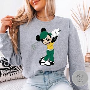 May include: A heather grey crewneck sweatshirt featuring a graphic print of Mickey Mouse dressed in a yellow shirt, green pants, and a green cap, holding a golf club. The character is depicted in a golf swing pose with white gloves and shoes.