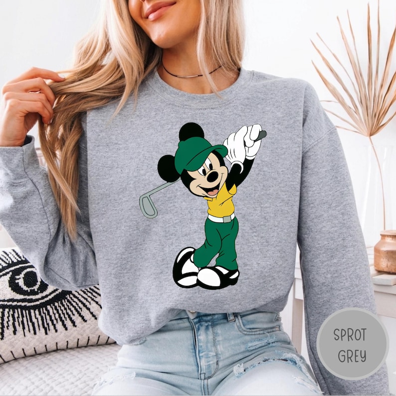 May include: A heather grey crewneck sweatshirt featuring a graphic print of Mickey Mouse dressed in a yellow shirt, green pants, and a green cap, holding a golf club. The character is depicted in a golf swing pose with white gloves and shoes.