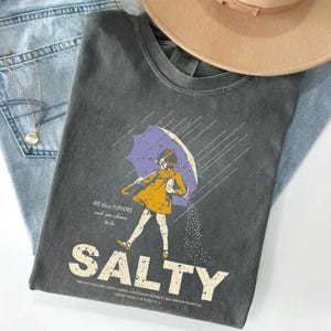 May include: Dark gray t-shirt with a graphic of a girl under a purple umbrella, with the word "SALTY" in large letters. The shirt also features the text "All these flavors and you choose to be". The shirt is paired with light blue jeans and a tan hat.
