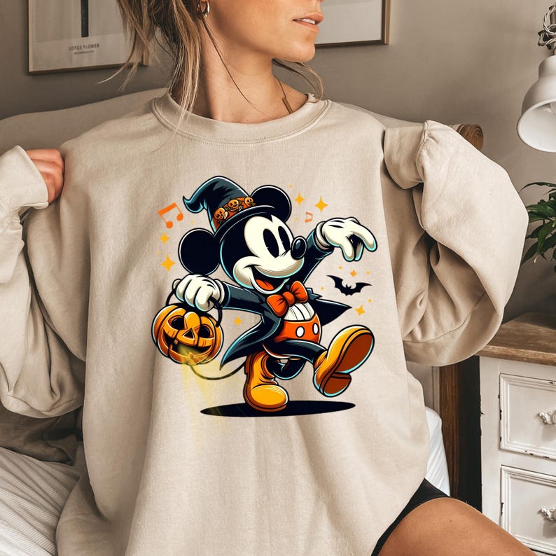 May include: A beige crewneck sweatshirt featuring a graphic of Mickey Mouse dressed in a black wizard hat with small orange pumpkin accents, a black tailcoat, and an orange bowtie. Mickey is holding an orange jack-o'-lantern bucket.