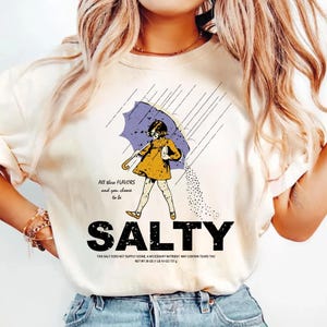 May include: Cream-colored t-shirt with a graphic of a girl under a purple umbrella, with the word "SALTY" in bold black letters. Text includes "All these flavors and you choose to be" and "NET WT 26 OZ (1 LB 10 OZ) 737 g".