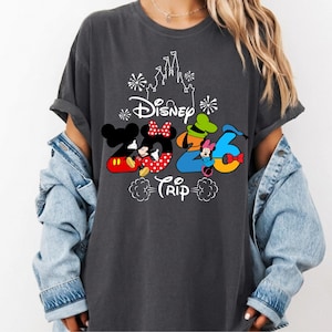 May include: A charcoal grey t-shirt featuring a white outline of a castle and fireworks above the text Disney 2026 Trip. The graphic includes colourful cartoon characters integrated into the numbers, with Mickey Mouse, Minnie Mouse, Goofy, and Donald Duck.