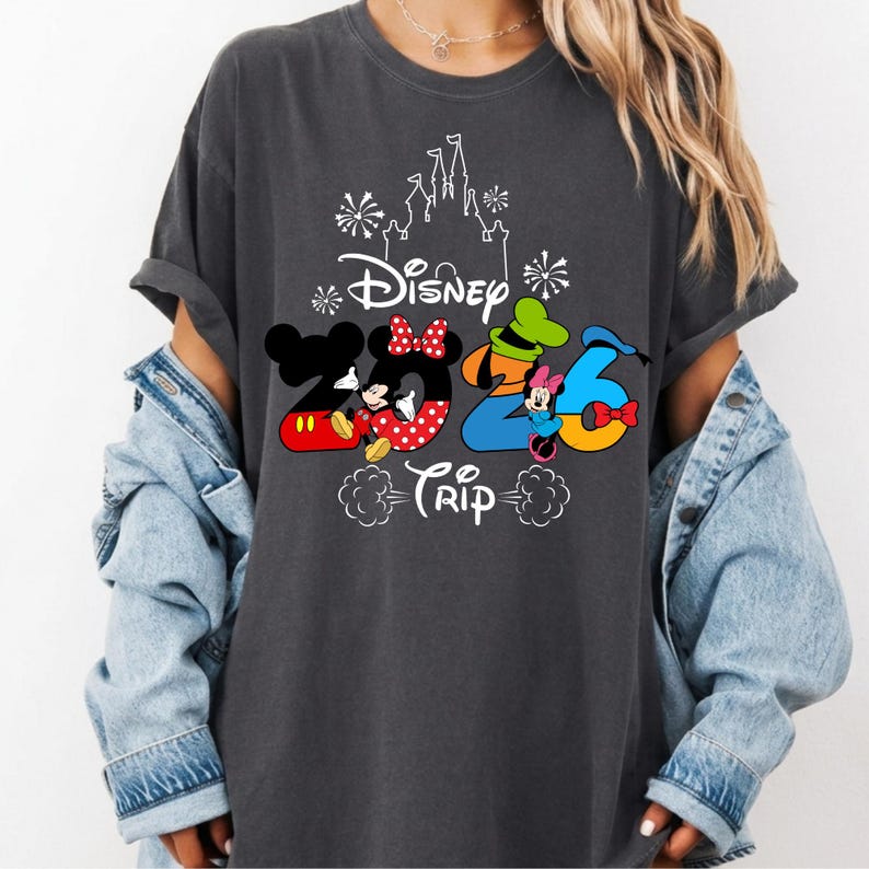 May include: A charcoal grey t-shirt featuring a white outline of a castle and fireworks above the text Disney 2026 Trip. The graphic includes colourful cartoon characters integrated into the numbers, with Mickey Mouse, Minnie Mouse, Goofy, and Donald Duck.