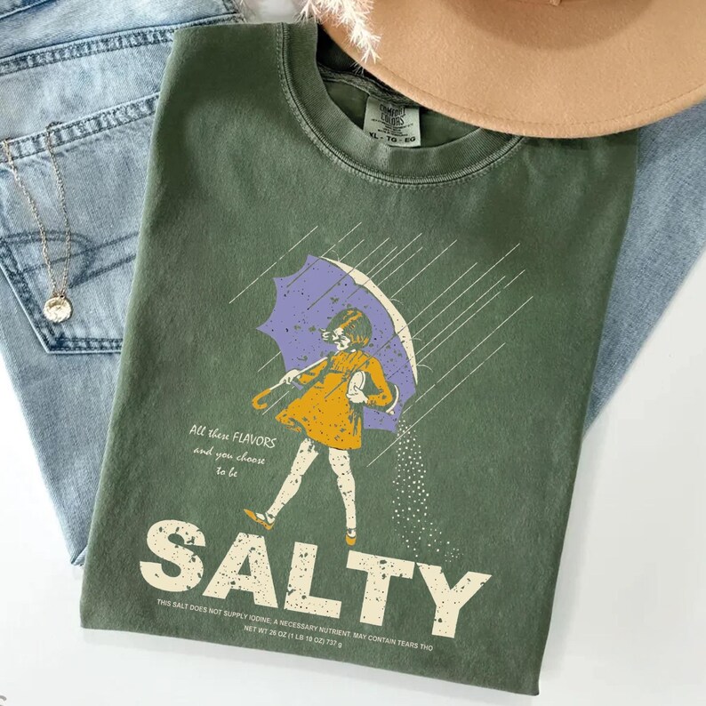May include: Olive green t-shirt with a graphic of a person under a purple umbrella, with the word "SALTY" in large white letters. The shirt also has text that reads "All these flavors and you choose to be".