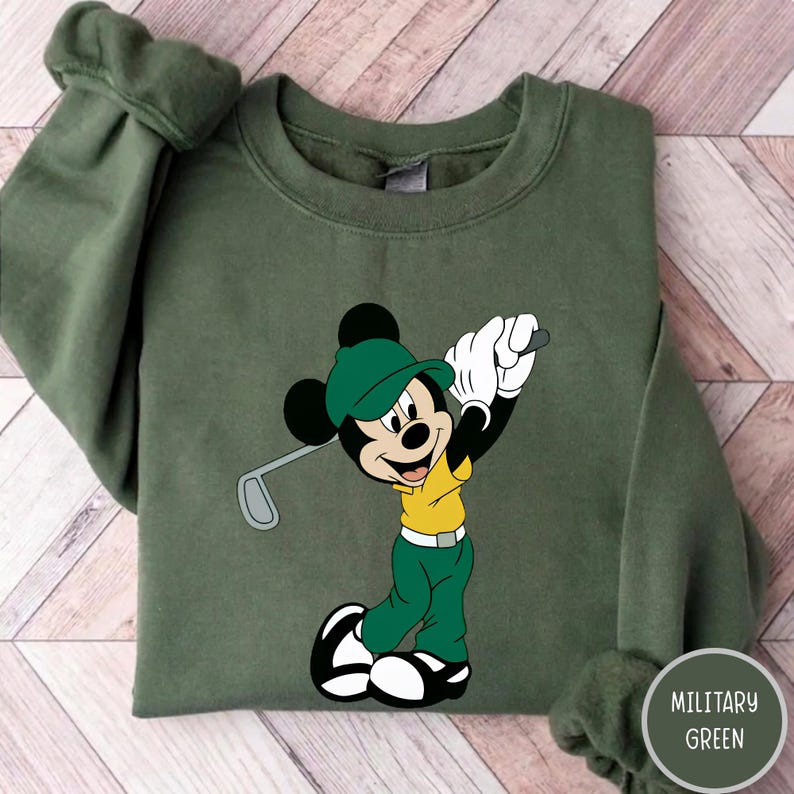 May include: A military green crewneck sweatshirt featuring a graphic of Mickey Mouse wearing a green cap, a yellow shirt, and green pants while holding a golf club. The character is depicted in a golf swing pose with white gloves and shoes.