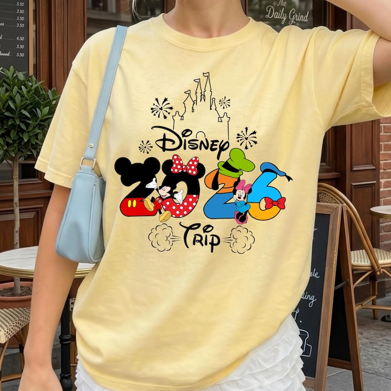 May include: A pale yellow short-sleeve t-shirt featuring a graphic print that includes the text Disney 2026 Trip, a castle outline, fireworks, and cartoon characters Mickey Mouse, Minnie Mouse, Goofy, and Donald Duck integrated into the numbers.