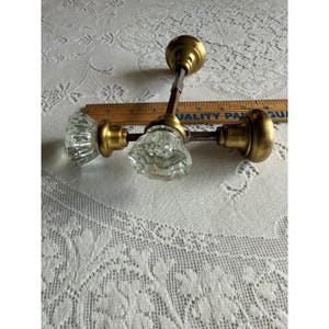 May include: Three door knobs with clear, faceted glass handles and brass-toned metal bases. Each knob is attached to a metal spindle. A wooden ruler is positioned behind the knobs, showing a measurement scale in inches along its edge.
