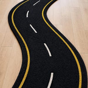 以下が含まれることがあります： 道路の形をした長く曲がりくねった黒いラグ。ラグの端には2本の連続した黄色の線があり、中央には白い破線が並んでおり、高速道路や車線の標示のような外観を模しています。