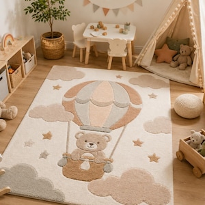 May include: A rectangular area rug featuring a central design of a teddy bear riding in a hot air balloon. The rug is cream-colored with textured, raised patterns of brown and gray clouds, stars, and the balloon, creating a soft, tactile surface.