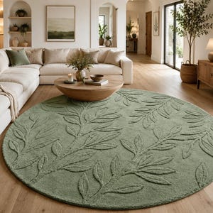 May include: A large, circular area rug in a soft sage green colour. The rug features a raised, textured botanical pattern of leafy branches that extend across the entire surface, creating a subtle, tactile design on the floor covering.
