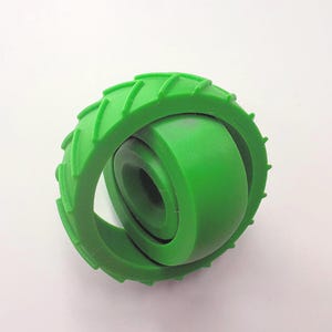 May include: A bright green, 3D-printed toy featuring a wheel-like design. The outer ring has a textured tread pattern, and the inner section is a solid, rounded shape. The toy is made of a smooth, solid material and is set against a white background.