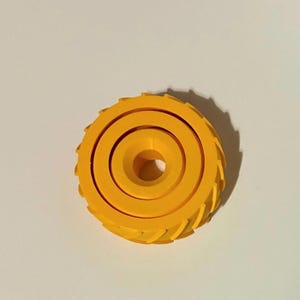 May include: A bright yellow, circular object with a central hole and concentric rings. The outer edge features a series of evenly spaced, triangular teeth. The object is set against a plain, off-white background.