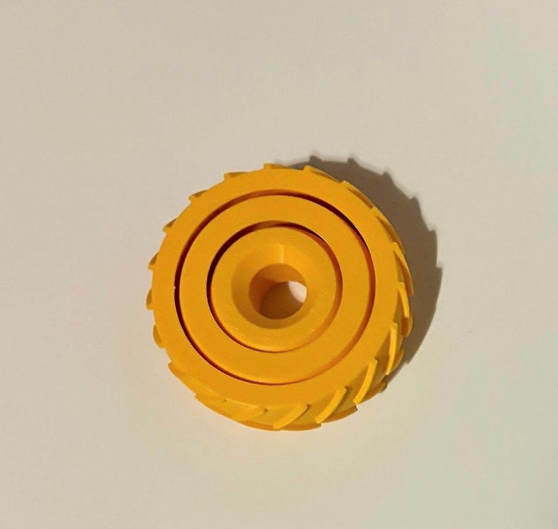 May include: A bright yellow, circular object with a central hole and concentric rings. The outer edge features a series of evenly spaced, triangular teeth. The object is set against a plain, off-white background.