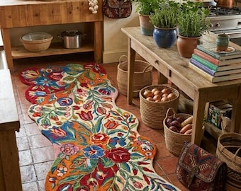 May include: A colorful, floral runner rug with a wavy edge. The rug features a vibrant mix of red, blue, orange, and green flowers and leaves on a cream background. The rug is placed on a terracotta tile floor in a kitchen setting.