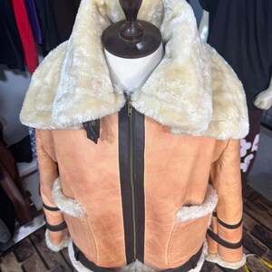 May include: A tan leather jacket with a cream-colored shearling collar and lining. The jacket features a zip-up front, brown trim, and pockets. The jacket is displayed on a mannequin.