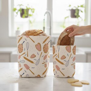 Peut inclure: Deux contenants à pain crème avec un motif répété d'outils de boulangerie et de formes roses et dorées. L'un contient un pain, et une main se tend vers l'autre pour en saisir un. Les contenants sont sur un comptoir blanc.