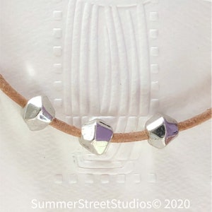 May include: A close-up of a brown leather cord with three silver faceted beads. The beads are geometric in shape and have a shiny finish.