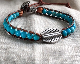 May include: A brown leather bracelet with turquoise beads and a silver leaf charm. The bracelet is tied with a knot and has a silver bead with a leaf design.