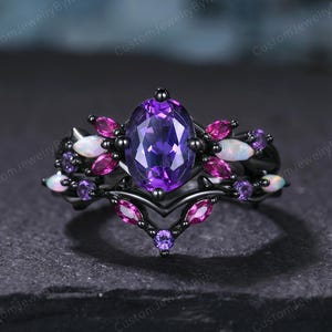 May include: A black ring with a large oval purple gemstone at the center, surrounded by smaller pink and white gemstones. The ring has a detailed, ornate design with a dark finish, showcasing a unique and elegant style.