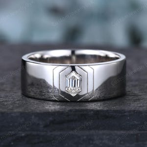 May include: A silver ring with a geometric design. The ring features a central, clear, rectangular-cut gemstone set within a hexagonal frame. The band is wide and polished, reflecting light. The ring is displayed on a dark, textured surface.