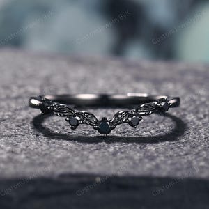 May include: A black, curved wedding band with a leaf design and black gemstones. The ring is set on a gray surface. The text "CustomJewelryByK" is visible in the background.
