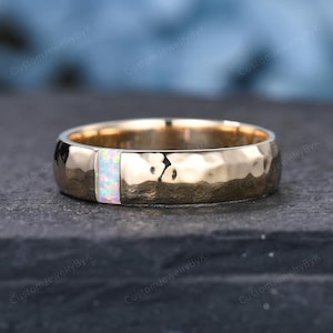 May include: A gold-colored ring with a hammered texture and a rectangular opal inlay. The opal features a mix of white, pink, and blue hues. The ring is set against a dark, textured surface.