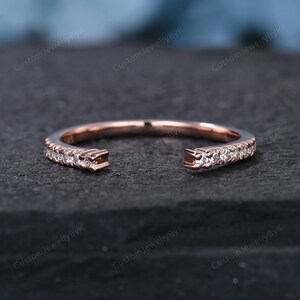 May include: Rose gold ring with a split shank design. The ring is encrusted with small, clear gemstones along the band. The open design features two squared-off ends. The ring is set against a dark, textured background.