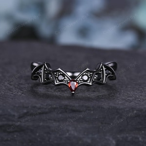 May include: A black ring with a bat wing design. The ring features black stones and a heart-shaped red stone at the center. The ring is set against a dark background.