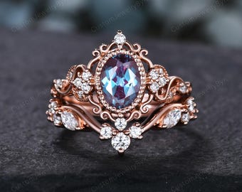 May include: Rose gold engagement ring set with an oval-cut, color-changing alexandrite gemstone. The ring is surrounded by small, round, and marquise-cut diamonds. The band features intricate filigree detailing, creating a vintage-inspired look.
