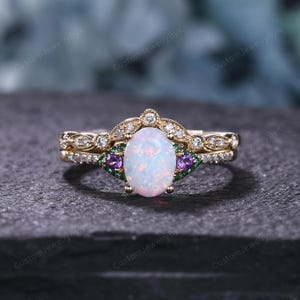 May include: A gold ring set featuring an oval opal center stone, flanked by purple and green gemstones. The band is adorned with small, clear gemstones. The ring is set against a dark, textured surface.