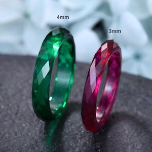 May include: Two faceted rings standing upright. The left ring is emerald green and labeled 4mm. The right ring is deep ruby red and labeled 3mm. Both rings feature a geometric, multi-faceted surface texture that reflects light across their edges.