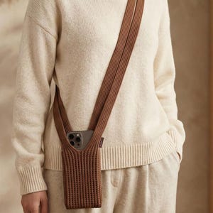 Może przedstawiać: Brązowe, dziergane etui na telefon z pasującym paskiem noszonym na ciele. Etui na telefon jest przeznaczone do przechowywania smartfona. Pasek jest wykonany z grubego, plecionego materiału. Tło jest w neutralnym kolorze.