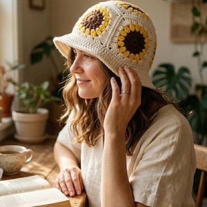 Könnte beinhalten: Cremefarbener Häkel-Eimerhut mit Sonnenblumenmustern in Gelb und Braun. Der Hut hat eine kleine Krempe. Der Hut ist aus weichem, strukturiertem Garn gefertigt. Eine Person trägt den Hut.