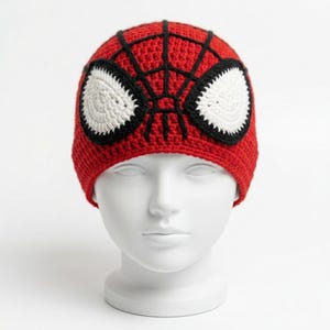 Pode incluir: Um gorro de crochê vermelho com um padrão de teia de aranha preto e dois grandes remendos ovais brancos para os olhos com bordas pretas. O gorro é exibido em uma cabeça de manequim branca, destacando a textura detalhada do fio.