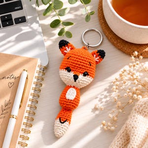 May include: A crochet fox keychain featuring a bright orange body, a white belly patch, and a white-tipped tail. The fox has black-tipped ears, black embroidered eyes, and a small black nose. A silver metal keyring is attached to the top of the head.