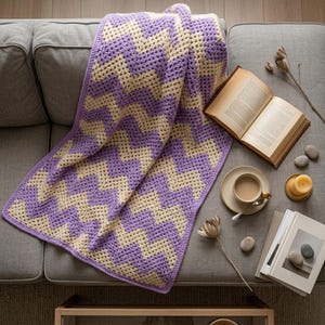 Puede incluir: Manta de ganchillo hecha a mano con un patrón de chevron, con filas alternas de lavanda y crema. La manta está sobre un sofá gris, creando una estética acogedora. Las dimensiones de la manta son de aproximadamente 127 cm x 152 cm.