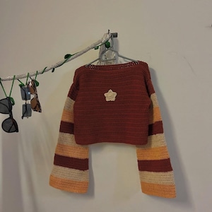 Könnte beinhalten: Ein gehäkelter, gecroppter Pullover in einem rötlich-braunen Farbton mit gestreiften Ärmeln in Creme, Orange und Braun. Auf der Vorderseite ist ein cremefarbener Stern aufgenäht. Der Pullover hängt an einem grauen Kleiderbügel vor einer weißen Wand.