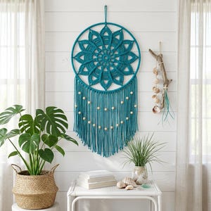 May include: A teal circular wall hanging featuring a crocheted mandala pattern inside a hoop, with long teal fringe strands adorned with light-colored wooden beads. The piece is suspended by a matching teal cord from the top of the circular frame.