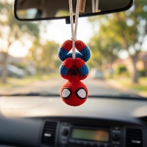 Op de afbeelding: Een gehaakt hangend ornament in de vorm van een rood en blauw figuur met een rond hoofd, grote witte ogen en een zwart spinnenembleem op de borst. Het figuur hangt ondersteboven aan een wit koord en is bedoeld voor gebruik in een voertuig.