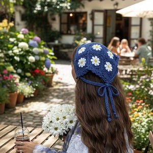 May include: A blue crochet head covering adorned with white daisy appliqués. The head covering is tied at the back with a matching blue tie. The person is holding a bouquet of white flowers and a beverage with a straw. The setting is an outdoor cafe.