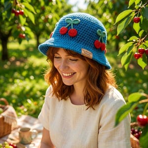 May include: A blue crochet bucket hat decorated with red cherry appliqués and green leaves. The hat has a small tag that reads "The Modern Pattern". The hat is worn outdoors in a cherry orchard.