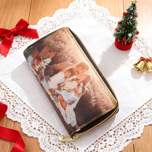 May include: A long, zippered wallet featuring a family portrait. The wallet has a gold-colored zipper and a black trim. The background includes a red bow, a small Christmas tree, and gold bells.