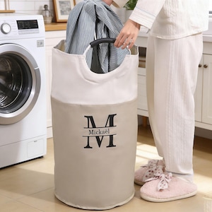 May include: A beige and white laundry hamper with a black handle and the name "Mickaël" printed on the front. The hamper is filled with folded blue and white striped clothing. A person is holding the handle of the hamper.