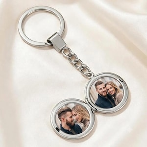 May include: A silver-toned metal keyring featuring a circular locket pendant. The locket opens to reveal two interior circular frames, each containing a photograph of a couple. The keyring includes a split ring and a short chain link connector.