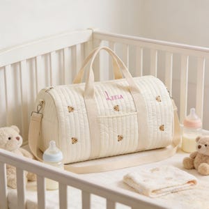 May include: Cream-colored quilted baby bag with embroidered teddy bear accents and the name "Leela" in pink. Features a shoulder strap and top handles. Includes a front pocket and is surrounded by baby items.