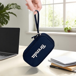 May include: A navy blue pencil case with a zipper closure and a wrist strap is held up by a hand. The name "Brenda" is printed in white script on the front of the case. The case is rectangular and appears to be made of fabric.