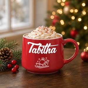 Op de afbeelding: Een rode keramische mok gevuld met warme chocolademelk, afgewerkt met slagroom en rode spikkels. De mok is voorzien van de witte tekst Tabitha in een scriptlettertype boven een witte lijntekening van een slee met een zak.