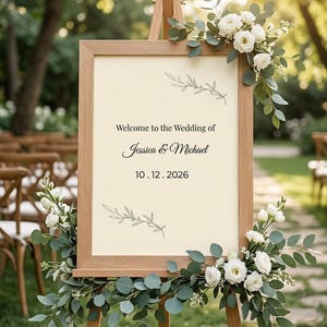 Puede incluir: Un caballete de madera clara que sostiene un letrero rectangular de papel color crema. El letrero presenta el texto "Welcome to the Wedding of Jessica & Michael" y "10.12.2026" con ilustraciones de hojas verdes. Flores blancas y eucalipto adornan el marco.