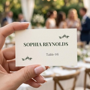 May include: A rectangular cream-colored place card featuring dark green text that reads SOPHIA REYNOLDS and Table 04. The card is decorated with small, dark green botanical illustrations of leafy branches positioned above and below the name.